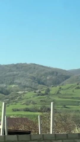 The video captures a scenic view from a moving vehicle, likely in Sayukovo, Russia, on a bright, sunny day with a clear blue sky. The landscape features lush green rolling hills, some covered with forests, leading up to distant mountains. Scattered houses with various roof types, including brown tiled and grey corrugated, are visible among the greenery. In the foreground, there are trees, some bare and some adorned with white blossoms, suggesting springtime. A patterned fence structure is briefly visible at the bottom of the frame. The scene conveys a sense of peaceful travel through a picturesque rural area. Muffled speech can be heard in the background.