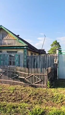 The video pans across the exterior of a weathered, wooden house with peeling paint and green shutters. A wooden fence, partially dilapidated, encloses the property. The yard is overgrown with grass and weeds, and a large tree is visible to the left of the house. The sky is clear and blue.