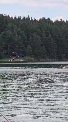 The video shows a body of water with a dense forest in the background. On the far shore, there is a campsite with several tents visible. In the water, there are two inflatable tubes or small boats. One is yellow and appears to be moving towards the left of the frame. The other is white and is further to the right, with a person visible in it. The water is rippled, indicating some movement. The sky is overcast.