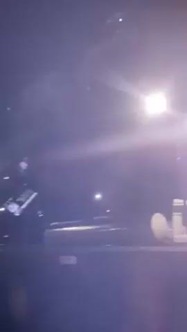 A tow truck is transporting a cylindrical object, possibly a tank, at night.  The scene is illuminated by bright lights, possibly from the truck itself or streetlights. The camera is low to the ground.