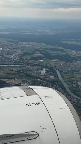 An aerial view from an airplane window captures a vast landscape. The wing of the plane, marked with "NO STEP" in black letters, partially obstructs the bottom of the frame. Below, a river winds through a densely populated area with numerous buildings and houses. Further into the distance, a large power plant stands prominently with multiple cooling towers and smokestacks. The surrounding terrain consists of green fields, trees, and what appear to be agricultural plots. The sky above is cloudy, and the overall lighting suggests either early morning or late afternoon.