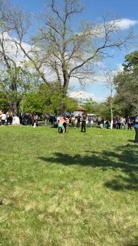 In a park-like setting on a sunny day, two men wrestle on a grassy lawn, surrounded by a crowd of onlookers. The man in green shorts ends up on top of the man in black shorts and appears to pin him down. The crowd consists of people of various ages and genders, some watching intently, others chatting among themselves. Trees provide shade, and tents are set up in the background, suggesting an organized event or gathering.
