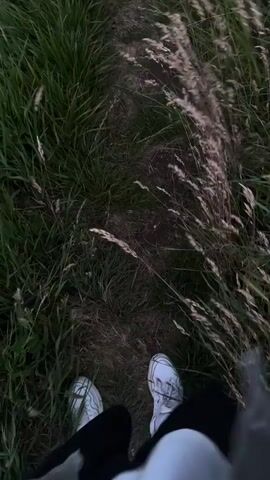 A low-angle shot shows a person's feet, clad in white sneakers and dark pants or shorts, standing on or walking along a narrow, worn dirt path. The path is surrounded by tall, lush green grass on the left, and a mix of green and drier, lighter-colored tall grass or reeds on the right. The overall impression is a natural, outdoor setting, possibly a trail or field, in Fécamp, France. The lighting suggests either an overcast day or dusk/dawn, as the colors are slightly muted. There is no visible text.