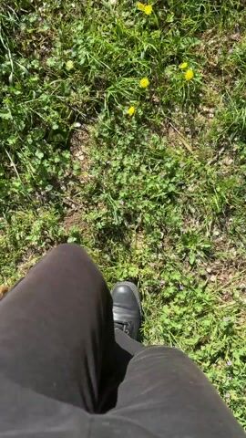 A first person view shows a person walking across a grassy area dotted with dandelions and arriving at the edge of a small waterfall. The person is wearing black shoes and brown pants. The waterfall is small and surrounded by green trees, dirt, and power lines are overhead. The sky is blue.