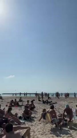 The video shows a beach scene on a sunny day. There are many people on the beach, some are playing Matkot, a beach paddle game, near the water, while others are sunbathing or relaxing under umbrellas. The beach is sandy, and the sky is clear blue. In the background, there are buildings and a pier. The camera pans across the scene, showing different groups of people and the overall atmosphere of the beach.