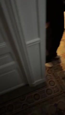 The camera moves from behind a door to show a group of people in a hallway. Some of the people are walking towards the camera, while others are standing around. One person is looking at their phone, another is wearing headphones, and another is wearing a burgundy jacket. The hallway has white walls, a wooden staircase, and pendant lights hanging from the ceiling.