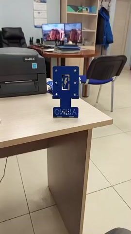 This media file depicts a close-up view of a desk in an office setting, likely in Selyatino, Russia. The primary focus is a blue laser-cut stand with a lightning bolt symbol and stars, placed on the desk in front of a printer. Below the lightning bolt, the Cyrillic word "ОЛИЦА" is visible. To the left of the stand is a black Godex printer. The desk itself is light wood-colored, and the surrounding floor is tiled in a light beige.

In the background, an office environment is discernible. There's a black leather office chair, two computer monitors displaying what appears to be landscape images, and a circular wooden table. A tall bookshelf unit contains various items, including what looks like a rolled-up mat and a blue jacket. Another chair, this one blue with a metal frame, is positioned near the table. The overall impression is a functional, well-lit office space. The time of day is not explicitly indicated but the lighting suggests it is daytime. There is no indication of weather conditions. The scene is static, with no discernible activity other than the static positioning of objects. The visible text on the stand is "ОЛИЦА".