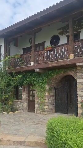 The video shows a traditional stone and wood building with a second-story balcony. The balcony has intricately carved wooden railings and is supported by thick wooden posts. The building's lower level features an arched entryway with ornate black metal gates, stone walls, and arched windows. Green vines are growing on the building's exterior, and there are various plants and flowers in the foreground, including bushes, yellow and orange flowers, and red roses. The sky is visible in the upper portion of the frame, appearing overcast. The video pans slightly from left to right, showcasing different sections of the building and its surroundings.