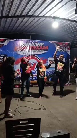 A group of people are gathered under a corrugated metal roof, possibly an outdoor event space. A large banner with the words "MUS CAMP MERDEKAAN" and "JATENG" is prominently displayed behind them. On the banner, there is a large "V" logo and images of mountains. A woman in a black dress stands to the left, facing the group. In the center, two women and one man are singing and dancing. One woman is wearing a red hijab and a black top, holding a microphone. The other woman is wearing an orange and black patterned scarf and an orange and black t-shirt. The man next to her is wearing a black t-shirt and jeans, and is raising his right arm in the air. To the far right, another person is partially visible. A cable runs across the floor in front of the performers. The ground is paved.