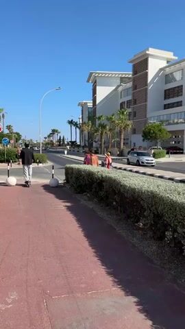 The video shows a street scene with modern buildings and palm trees. There is a red paved walkway on the left side of the screen. A person on a skateboard is moving away from the camera on the walkway. Across the street, there are several people near a crosswalk. Some are standing and talking, while others appear to be walking. There are cars on the street, including a silver sedan and a white SUV. A stop sign is visible on a pole. The sky is clear and blue. The overall impression is of a sunny day in a resort town.