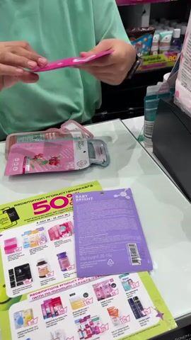 A person wearing a light teal shirt is standing behind a counter, holding a pink package with text and images on it. Their right arm is extended, and their hand is touching the package. In front of them on the counter, there are several items including a pink pouch with text and images, a purple pouch with text and images, and a flyer with various product images and text. The flyer has the words "REDEMPTION PRODUCT" prominently displayed. To the right of the counter, a black cash register is partially visible. The background shows shelves with various products. The lighting appears to be artificial and bright.