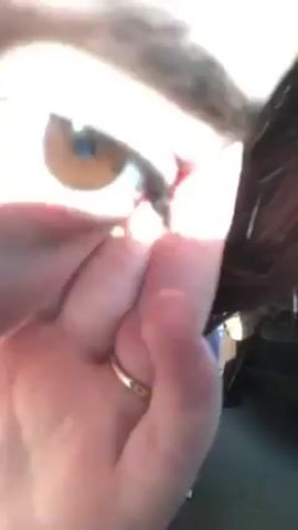 A close-up shot shows a person's eye, brown in color, partly obscured by their fingers. The person is wearing a gold ring. Light is shining brightly on the eye and hand. The background is dark, making it difficult to discern the surrounding environment. Some strands of hair are visible near the eye.