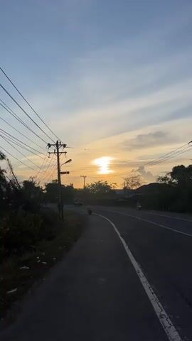 A vertical video shows a road at sunset. The sun is low on the horizon, casting a warm glow across the sky. The road is paved and curves to the right. There are telephone poles and power lines on the left side of the road. Trees and vegetation are visible on both sides of the road. A few vehicles, including cars and motorcycles, are seen on the road. The sky is a mix of blue and orange hues, with some clouds. The overall atmosphere is peaceful and serene.
