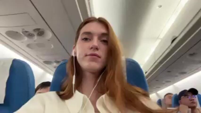 A woman with long red hair wearing a white shirt and earbuds is in the foreground, seated in a blue airplane seat. The airplane cabin is filled with passengers, most of whom are looking forward. An airplane staff member stands in the aisle in the middle of the cabin. The cabin is lit by overhead lights. The overhead compartments are visible. The video is shot from the seated perspective of the woman in the foreground.