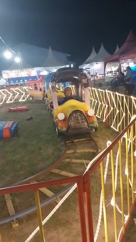 The video shows a child riding on a colorful amusement park train. The child is wearing a yellow hooded jacket and is looking forward. The train has multiple cars, each with a different color scheme. The train is on a track that is lined with lights. In the background, there are tents and other amusement park attractions. The scene appears to be at night, as evidenced by the lighting.