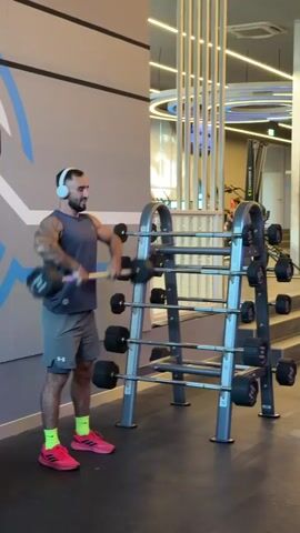 The video shows a man with tattoos on his arms and a beard, wearing a grey tank top, grey shorts, yellow socks, and pink athletic shoes. He is also wearing white headphones. He is performing a upright row exercise with a barbell. The barbell has black weight plates on either end. The man is standing in a gym, with a rack of barbells behind him. The gym appears to be modern and well-lit, with a grey wall on the left featuring a blue and white abstract design. The floor is dark. In the background, there are various gym equipment and reflective surfaces. The lighting suggests it is daytime. The man appears focused and determined, with no discernible expression of emotion. The time of day is not specified, but the lighting indicates it is not night.