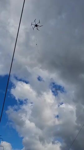 A drone is suspended in the air by a tether, which is connected to a large spool of cable on the ground. A person wearing a high-visibility vest is operating a remote control, presumably to maneuver the drone. The scene is set in an open field with some trees and buildings in the background. The sky is cloudy.