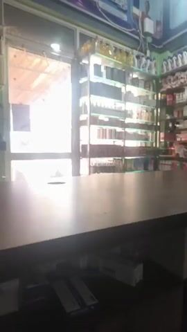 The video shows a man in a blue shirt sitting in a pharmacy. He is lighting a cigarette with a lighter. Shelves filled with medicine boxes are visible in the background.