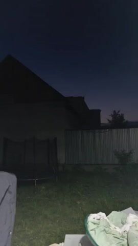 The video is of a backyard at dusk. A trampoline is visible on the left side of the frame. A house is in the background with a corrugated metal fence. The sky is a deep blue.