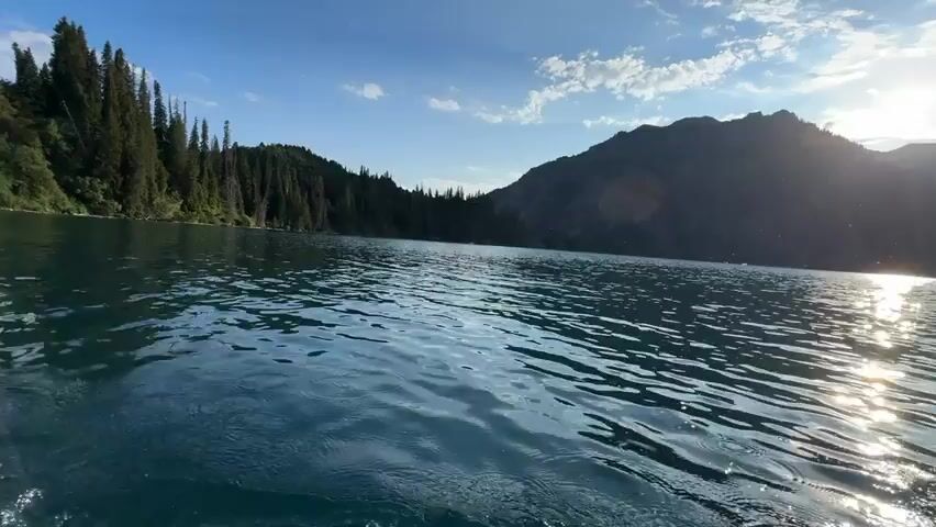 This media file depicts a scenic view of a lake in Arkit, Kyrgyzstan. The camera is positioned low to the water, suggesting it might be mounted on a boat or a person in the water. The foreground is dominated by the turbulent, foamy wake of a moving object, likely a boat, disturbing the otherwise calm, clear blue water.

In the background, a dense forest of tall, dark green coniferous trees lines the left side of the lake, climbing up a steep mountainside. The right side of the frame is framed by a majestic, rugged mountain range, its peaks silhouetted against the sky. The sun is visible as a bright, slightly diffused orb high in the sky, casting a glittering reflection on the water's surface and creating lens flares.

The sky above is a clear, bright blue, dotted with scattered white, fluffy clouds. The overall impression is one of a beautiful, serene natural landscape during what appears to be a sunny day, possibly late afternoon given the angle of the sun and the quality of light. There are no people or man-made objects other than the implied boat visible in the frame. The mood evoked is one of adventure and immersion in nature, with the dynamic water suggesting movement and exploration. No visible text is present in the media file.