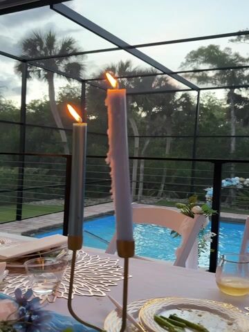 The video captures an outdoor dining scene at dusk or evening, likely on a screened-in patio overlooking a pool. The setting suggests a warm, tropical climate, reinforced by visible palm trees and lush foliage in the background.

The focus is on a beautifully set dinner table, adorned with a blue and white theme. Two lit candles in gold holders, one heavily melted, provide a warm glow. Place settings include decorative chargers, white plates, wine glasses, and a blue table runner. Floral arrangements in white and blue also decorate the table.

Several people are visible enjoying their meal. One woman, wearing a blue top, is seen eating from her plate with a fork. Other diners, including a man in a white shirt, are partially visible in the background, engaged in the dining experience. A can labeled "Topo Chico" is visible on the table.

Beyond the table, a sparkling blue swimming pool is prominently featured. White chairs with floral decorations are arranged around the pool area, hinting at additional seating or a decorative setup. The overall atmosphere appears relaxed and festive, indicating a pleasant dinner party or gathering. The ambient light suggests the transition from day to night, creating a cozy and inviting ambiance for the outdoor meal.