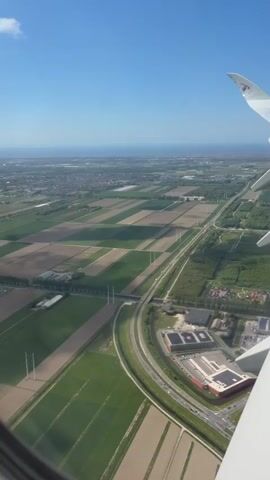An aerial view from an airplane window shows a landscape consisting of fields, roads, and buildings under a clear, blue sky. The fields are arranged in a patchwork pattern of light and dark green, possibly indicating different crops or stages of growth. A highway curves through the landscape, with cars visible on its surface. Nearby, a cluster of buildings, possibly a business park or industrial area, is seen. In the foreground, the wing of the airplane is visible, indicating the perspective from inside the aircraft. The overall scene suggests a rural or suburban area with agricultural and commercial elements.