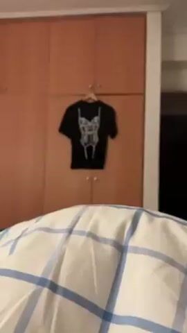 The video shows a black t-shirt with a printed corset design hanging on a wooden hanger. The t-shirt is displayed against a backdrop of a light brown wooden closet with small, round silver knobs. In the foreground, a white and blue plaid blanket is visible, suggesting a bed or seating area. The camera focuses on the t-shirt, gradually zooming in to highlight the corset print. The environment appears to be a bedroom or living space.