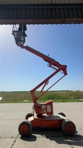 The video depicts a man working on a raised aerial work platform, also known as a cherry picker, under an overhang. The cherry picker is orange and black and stands on a paved surface outdoors. The man is wearing a black shirt and appears to be doing some kind of work on the underside of the overhang. The sky is clear and blue, and in the background, there is a grassy area and wind turbines in the distance. The camera remains stationary throughout the video.