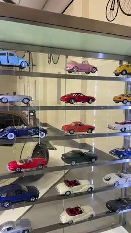 A camera pans slowly to the right across a glass display cabinet containing several shelves full of model cars of various makes, colors, and ages. A bicycle is mounted on the wall above the cabinet. The light in the room is bright. Some reflections can be seen in the cabinet glass.