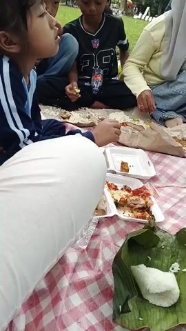 A group of people are having a picnic on a checkered mat. The camera is positioned at a low angle, looking across the picnic spread. In the foreground, there is a container with rice wrapped in banana leaves and another container with cooked noodles. Further back, there are white disposable food containers with remnants of food, including what appears to be chicken and sauce, and some loose rice. A person in white pants and a blue and white jacket is in the left foreground, holding food. To their right, another person, partially visible, is also eating. In the middle ground, a person wearing a black shirt with white lettering is seated. A bottle of water is visible. To the right, a person in a light yellow top and a blue headscarf is seated, holding a smartphone. Another person in a tan headscarf is visible further to the right, and a small child in a pink tutu is also present. The background shows greenery, suggesting an outdoor setting like a park. The overall scene depicts a casual meal shared outdoors.
