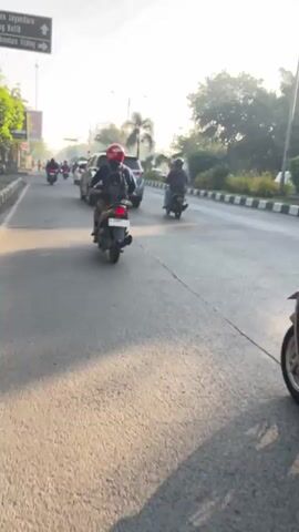 The video shows a view from a vehicle moving forward on a paved road, likely a street in a city. The road is bordered by sidewalks and buildings on the left, and greenery, trees, and a curb on the right. Several motorcycles and scooters are also on the road, moving in the same direction. The vehicles are diverse in type and color, with some riders wearing helmets of various colors, including black, red, and blue. The time of day appears to be early morning or late afternoon, given the angle of the sun and the shadows cast on the road, suggesting a well-lit environment. The atmosphere seems to be active with traffic, but not overly congested. The video is recorded from the perspective of someone inside a vehicle, capturing the motion and the surrounding environment as they travel. The overall impression is one of everyday urban transit.