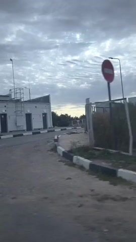 The video shows a gas station with a building and a canopy over the pumps. The sky is cloudy and the lighting suggests it is early morning or late evening. The camera appears to be moving, possibly from inside a vehicle. There are several signs and utility equipment visible at the gas station.