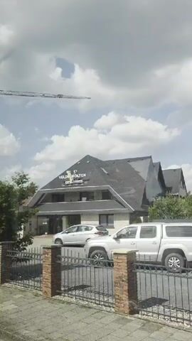 A camera pans from left to right across a building that appears to be a business or office. The building has a dark roof and light-colored walls. A sign on the roof reads "VELDSCHOLTEN." There is a fence in the foreground with brick posts and metal railings. A silver car is parked in front of the building, and part of another car is visible to the right. Trees and greenery are present on the left and right sides of the frame. The sky is cloudy. A crane is visible in the upper left corner of the frame. The video is approximately 5 seconds long.
