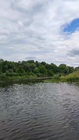 This is a landscape video of a wide river with calm water. The river is bordered on the left by a dense line of green trees and foliage, with a slightly hilly terrain visible further back. The sky is partly cloudy with patches of blue. In the far right of the frame, a red and white striped pole with a white circular sign is visible on the bank, possibly indicating a marker or a navigation aid. The video appears to be filmed from a moving boat or vessel.