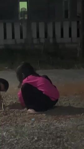 A young child with dark hair, wearing a bright pink long-sleeved shirt and black pants, is crouched down on a surface of gravel and dirt. The child is facing away from the camera and appears to be digging or playing with the ground. There is a hint of another person, likely another child, to the left of the frame, also partially visible. The background suggests an outdoor setting at dusk or night, with a building or structure with illuminated windows and a fence visible. The overall lighting is dim.