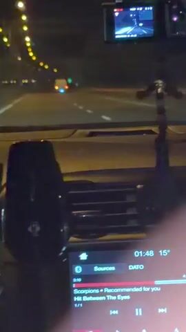 The video is filmed from the perspective of someone driving at night. The dashboard of a car is visible in the foreground, with a touch screen displaying music information. The song playing is "Hit Between The Eyes" by Scorpions. Outside the car, a highway is visible with streetlights lining the left side of the road. Other vehicles, including a truck and a car, can be seen on the road ahead. A dashcam is mounted on the windshield, showing a live feed of the road. A decorative item, possibly a car air freshener, is hanging from the rearview mirror. The overall atmosphere is that of a nighttime drive.