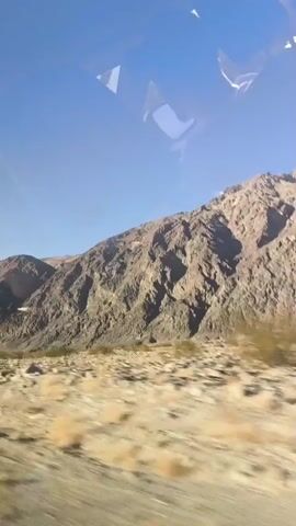 The video shows a view from a moving vehicle, likely a car, traveling through a desert landscape. The terrain is arid with sparse, dry vegetation and sandy soil. In the background, large, rocky mountains dominate the horizon under a clear blue sky with a few scattered clouds. The sun appears to be shining, casting shadows and highlighting the textures of the mountains and the ground. The camera seems to be positioned to capture a side view of the passing scenery, with reflections visible on the window.