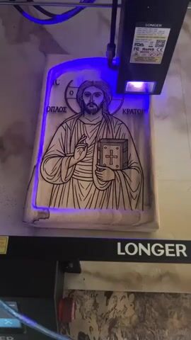 A laser engraving machine is shown in operation, engraving an image of Jesus onto a wooden plaque. The laser beam, emitting a blue light, moves across the surface of the wood, creating intricate lines and details. The plaque is rectangular in shape with rounded corners and features the image of Jesus in a traditional iconographic style, holding a book and raising his hand in a blessing gesture. Greek text is visible around the halo of Jesus. The machine's laser module is mounted on an arm that moves in a coordinated fashion to follow the design. The surrounding environment appears to be a workshop or a dedicated engraving space, with other equipment and materials visible in the background. The overall scene depicts the process of creating a custom wooden engraving using advanced technology.
