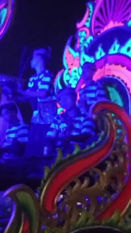This video shows a group of people in striped clothing standing on a platform decorated with neon lights in pink, green, and blue. The platform appears to be part of a parade float, with ornate, carved elements in red and gold visible in the foreground. The lighting is dark, with the neon lights providing the primary illumination. The people are looking in different directions and some appear to be holding objects, though their specific actions are not clear due to the low light and movement. There are multiple people on the platform, some standing and some appearing to be seated or crouched. The overall scene suggests a nighttime festival or performance with a vibrant, illuminated aesthetic.