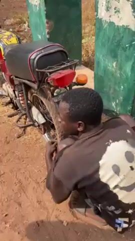 The video shows a man squatting next to a red and black motorcycle, apparently working on the rear tire. The motorcycle is parked on a dirt surface next to a green and white post. In the background, several people are sitting or standing under a thatched roof structure. Another man in a red shirt and black pants walks across the scene. The setting appears to be a rural area.
