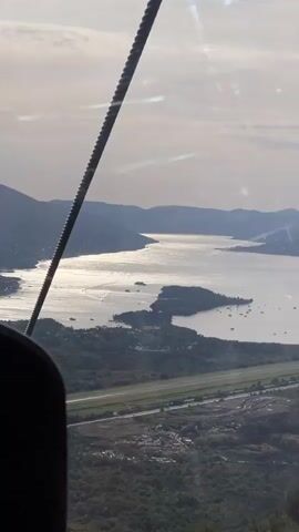 The video presents an aerial view captured from a moving vehicle, evidenced by a dark, ribbed cable running diagonally across the upper left and a dark, out-of-focus object in the bottom left corner, likely part of the vehicle's interior. Through a window with subtle reflections, a wide landscape unfolds, featuring a prominent airport runway with two parallel strips, situated on green land with some cleared or disturbed areas. Beyond the runway, a large body of water, possibly a bay or fjord, stretches into the distance, dotted with numerous small islands and boats. The water reflects bright sunlight, creating a shimmering effect. Distant mountains rise on either side of the waterway, and the sky above is bright and hazy.
