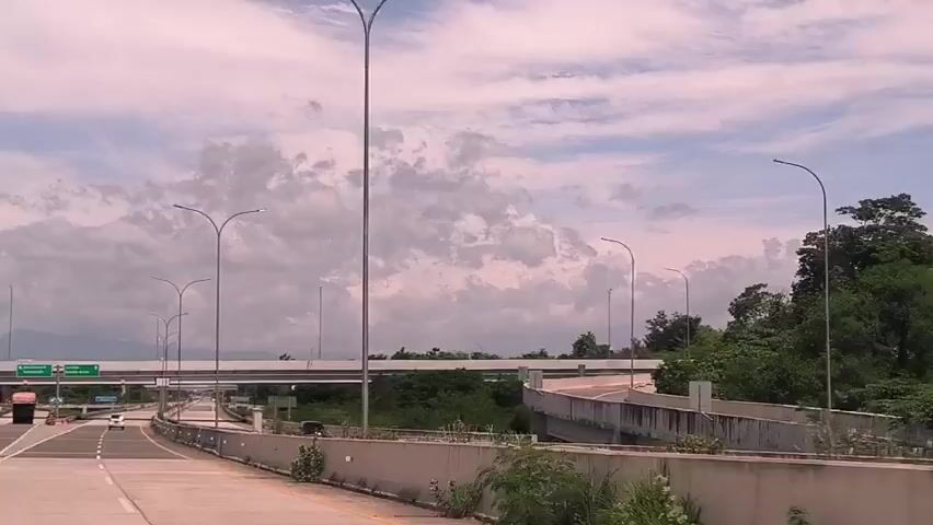 The video shows a view from a vehicle driving on a highway. The road has multiple lanes separated by dashed white lines. Concrete barriers line the edges of the road.  Tall streetlights are spaced along the highway. An overpass is visible ahead. There are a few vehicles visible on the road, including a truck and a car. The sky is cloudy. There is some greenery on the sides of the road. A sign indicates directions to "Bandara Int'l" and "Banda Aceh."  There is a yellow diamond sign on the left side of the road.