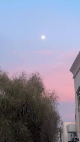 The video shows a view of the sky with a full moon. There is a large tree in the foreground, and a white building is visible on the right side of the frame. The sky is a blend of blue and pink, creating a sunset effect. The sound of a religious chant is audible.