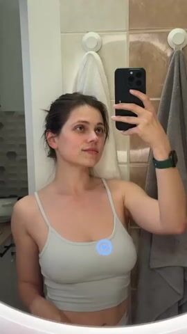 A young woman is taking a selfie in a bathroom. She is wearing a light gray tank top and has her hair in a messy bun. She is holding a black phone in her right hand and looking at the screen. On her left wrist is a green smartwatch. There is a round mirror in front of her. Behind her are beige wall tiles, a white towel hanging on a hook, and a gray towel hanging on a hook. In the lower left corner is a glimpse of a kitchen area with gray hexagonal tile backsplash. A blue circle with an arrow inside is digitally superimposed onto her chest in the center of the frame. She touches her left ear with her left hand.