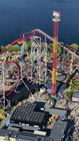 An aerial drone shot shows an amusement park with a large roller coaster and a drop tower ride. The roller coaster features red tracks and white supports, with several cars visible on the track. The drop tower, labeled "Boom," is tall and red and yellow, with a single car at the top. People are gathered around the rides and walkways of the park. The park is situated next to a body of water. The sun is shining, casting shadows and illuminating the scene.