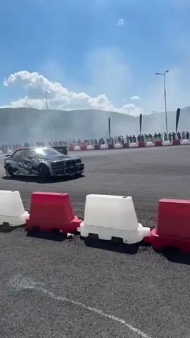 The video shows a drift racing event with cars on a track. In the foreground, a line of white and red plastic barriers are visible. In the background, a black and silver drift car with the name "PENEV" on its side is shown sideways on the track, producing smoke from its tires. Spectators are gathered behind a fence in the background. The sky is blue with some clouds, and there are mountains and trees in the distance. The event appears to be taking place outdoors on a sunny day.