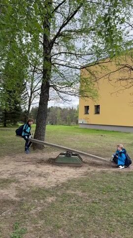 Two children are on a seesaw in a park-like area. One child is wearing a blue and black tracksuit and a backpack, while the other is wearing a blue jacket and a backpack. They take turns going up and down on the seesaw. A tree is next to the seesaw, and a yellow building is in the background. The ground is covered with dirt, grass, and some patchy green areas.