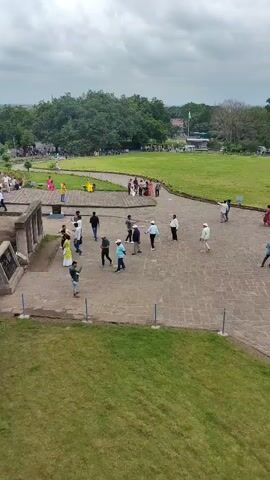 The video shows a group of people walking around a large, stone-paved courtyard in front of a massive, carved rock structure. The rock structure appears to be an ancient temple or monument, with intricate carvings and doorways. There are several people scattered throughout the courtyard, some walking individually and others in small groups. A grassy area is visible beyond the courtyard, with trees in the background. The sky is overcast, suggesting a cloudy day. One section of the rock face shows signs of excavation or construction, with scaffolding visible.