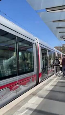 A modern tram waits at an outdoor station on a sunny day. Several people, mostly men, stand on the platform, waiting to board or having just exited the tram. The tram is painted silver with a broad red stripe and etched images of city scenes near the bottom. The station has a transparent roof for shelter. The scene is brightly lit.