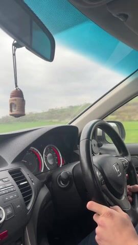 The video, shot from inside a car, shows a person driving on a rural road on a cloudy day. The car's dashboard, featuring a stereo system and instrument panel with red accents, is prominent. An air freshener hangs from the rearview mirror. The road ahead stretches between green fields under a gray, overcast sky. Other vehicles are visible in the distance. Raindrops are splattered on the windshield. The driver’s hand is visible on the steering wheel.